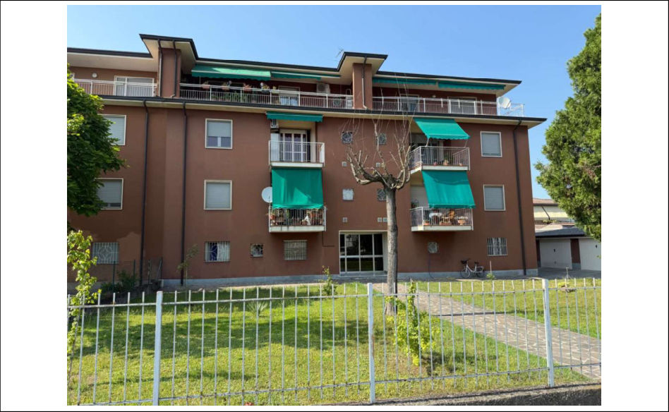 Immagine di un edificio residenziale pluripiano con recinzione e area verde circostante, ripresa da prospettiva esterna frontale.